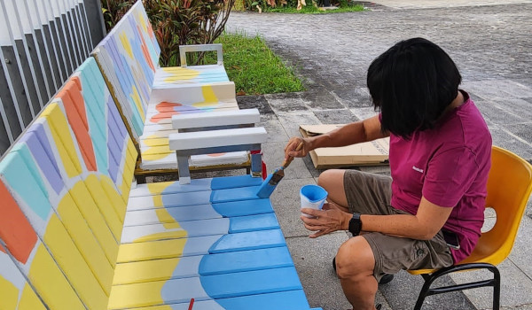 A person paints a colorful bench outdoors. They sit on a yellow chair and use a brush and blue paint.